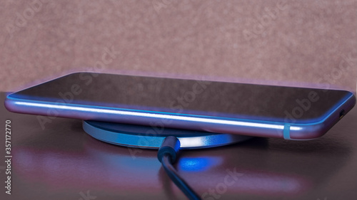 Charging smartphone on a round wireless charger with blue lights and luminous lines on a brown rough background and glossy surface
