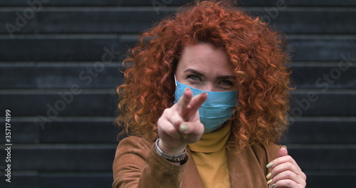 Beautiful young red-haired caucasian woman in medical mask looking into the camera touch hair