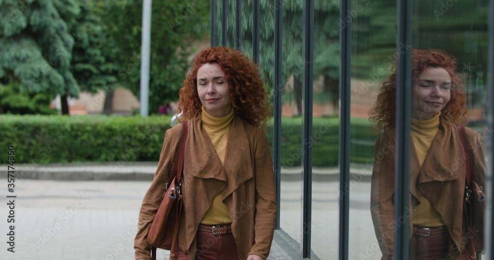 Fototapeta premium Attractive young red-haired curly woman with a bag on her shoulder walks near the glass wall looking at the camera