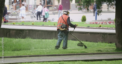 Worker in orange overalls mows the grass on the lawn with a gasoline mower, slow motion