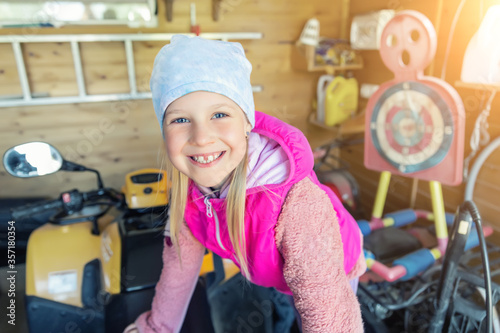 фотографія Cute adorable playful caucasian little happy blond girl portrait enjoy having fun playing at dad atv garage warehouse at home backyard