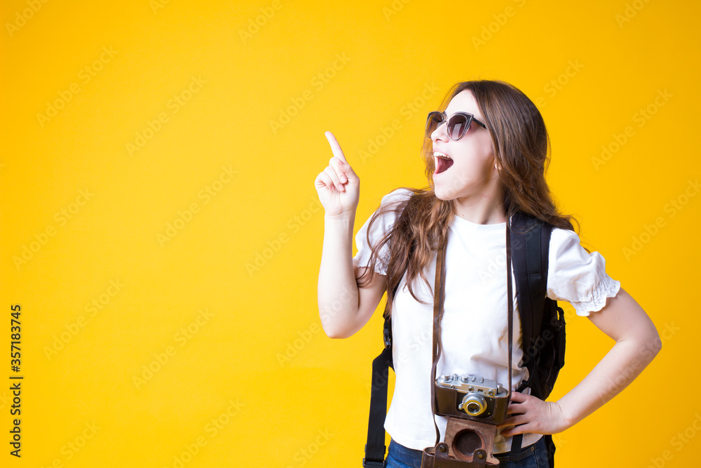 Excited tourist girl pointing finger away on blank copy space for your advertising.Pretty young woman traveler in white t-shirt,backpack,sunglasses and retro photo camera isolated on yellow background Excited tourist girl pointing finger away on blank copy space for your advertising.Pretty young woman traveler in white t-shirt,backpack,sunglasses and retro photo camera isolated on yellow background