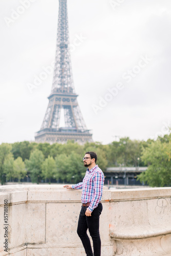 Handsome bearded young man in glasses and casual checkered shirt enjoying wonderful view of Paris and Seine river, standing on the bridge and looking away. Eiffel tower on the background © sofiko14