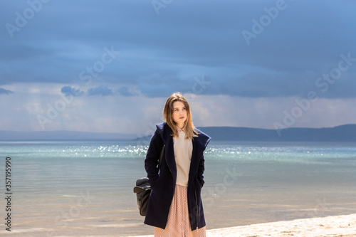 Fototapeta Naklejka Na Ścianę i Meble -  A young adult beautiful girl stands on the shore against the backdrop of an approaching cloud with rain. The woman is wearing a spotty dress, a white sweater and a navy coat.