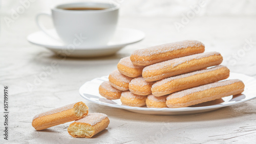 Traditional savoiardi biscuits or ladyfingers cookies on a plate and white cup of coffee on background. Selective focus