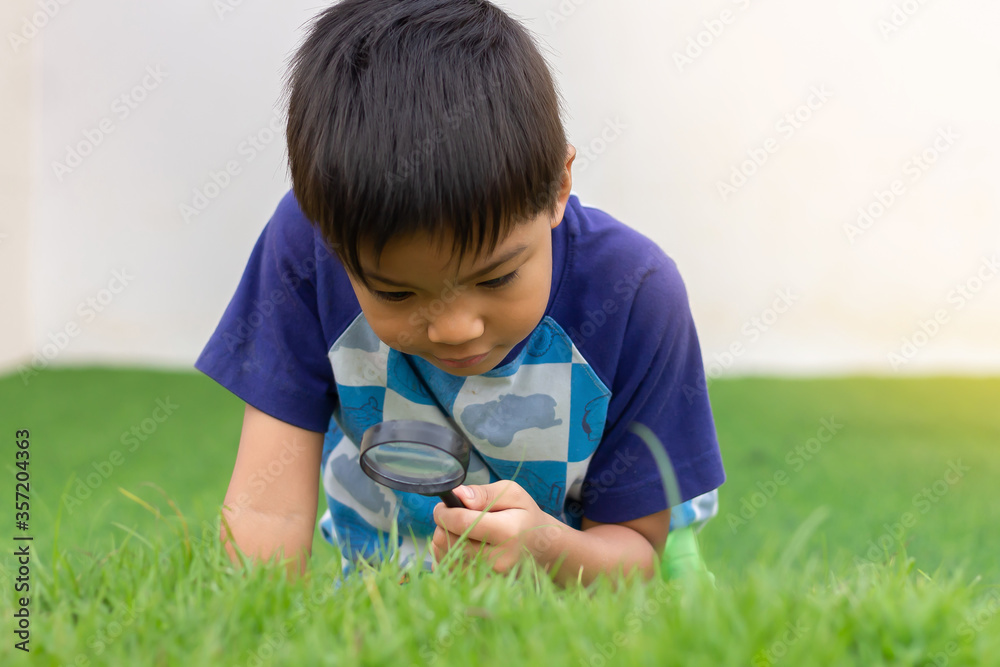 Portrait of happy Asian child boy holding and looking with magnifying glass on the flower tree and green grass field floor. Adventure, explorer and learning kid. Childhood 5-6 years old.