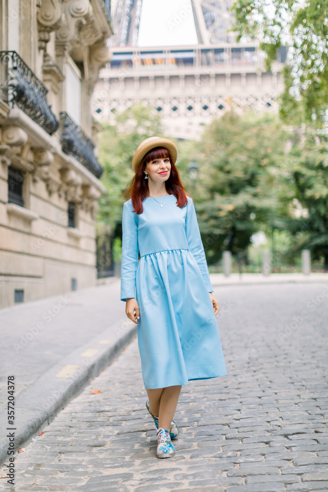 Street view of pretty young red haired lady, wearing elegant blue dress and straw hat, walking during the morning in Paris, posing to camera. Eiffel tower behind the woman