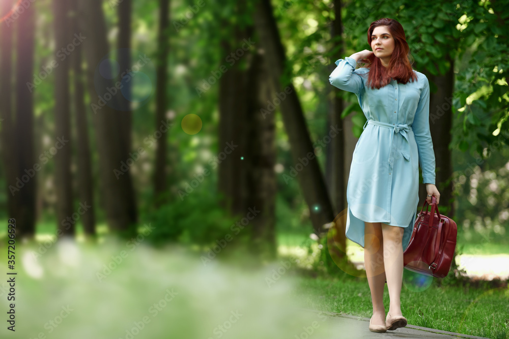 a young beautiful girl of Caucasian race in a light blue dress and a red bag in her hands is walking along a path in a green forest. a woman walks in nature and adjusts her red hair with her hand a young beautiful girl of Caucasian race in a light blue dress and a red bag in her hands is walking along a path in a green forest. a woman walks in nature and adjusts her red hair with her hand