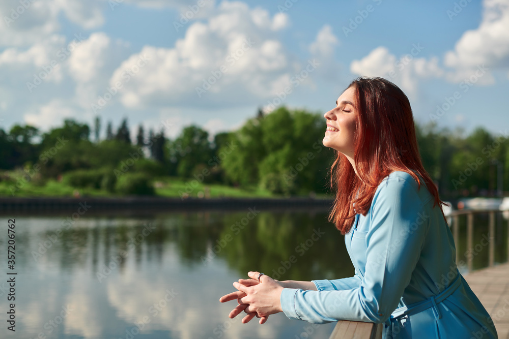 a beautiful young girl stands in nature and looks at the sun with her eyes closed. a girl with red hair enjoys summer and warmth