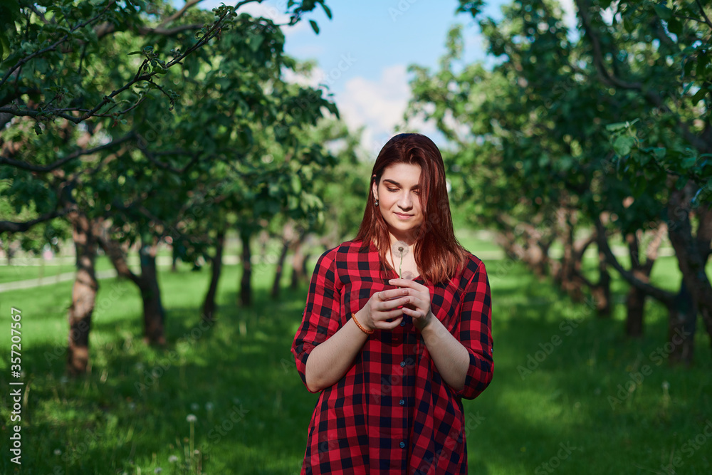 Fototapeta premium a young girl in a black and red checked shirt with red hair stands on a natural green background in a Park and holds a white dandelion in her hands, looks at it and smiles