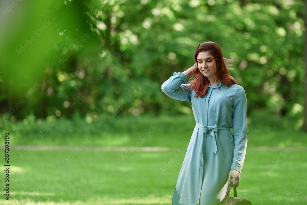 a young beautiful girl of Caucasian race in a light blue dress and a red bag in her hands is walking along a path in a green forest. a woman walks in nature and adjusts her red hair with her hand a young beautiful girl of Caucasian race in a light blue dress and a red bag in her hands is walking along a path in a green forest. a woman walks in nature and adjusts her red hair with her hand