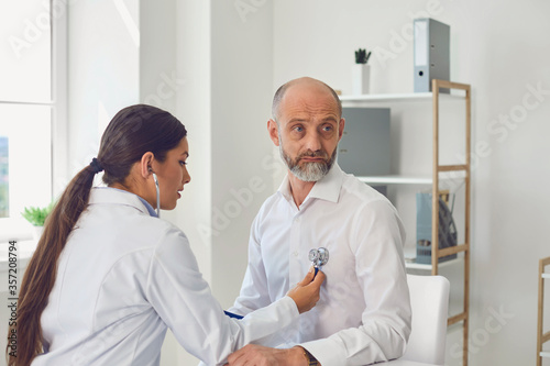 Woman doctor cardiologist listens with stethoscope to patient senor man in clinic office. Visit to the doctor in a medical hospital.