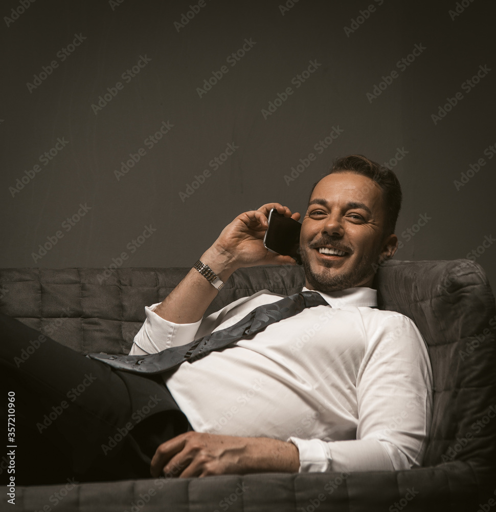 Fototapeta premium Businessman talks over mobile phone lying on sofa. Smiling man at couch talking smartphone on grey back. Close up shot. Toned image.