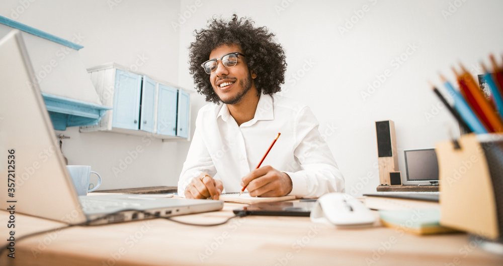 Happy millennial work at home workplace. Shaggy young man takes notes while looking at a computer monitor sitting at table studio apartment. Distance learning or home education concept. Toned image. Happy millennial work at home workplace. Shaggy young man takes notes while looking at a computer monitor sitting at table studio apartment. Distance learning or home education concept. Toned image.