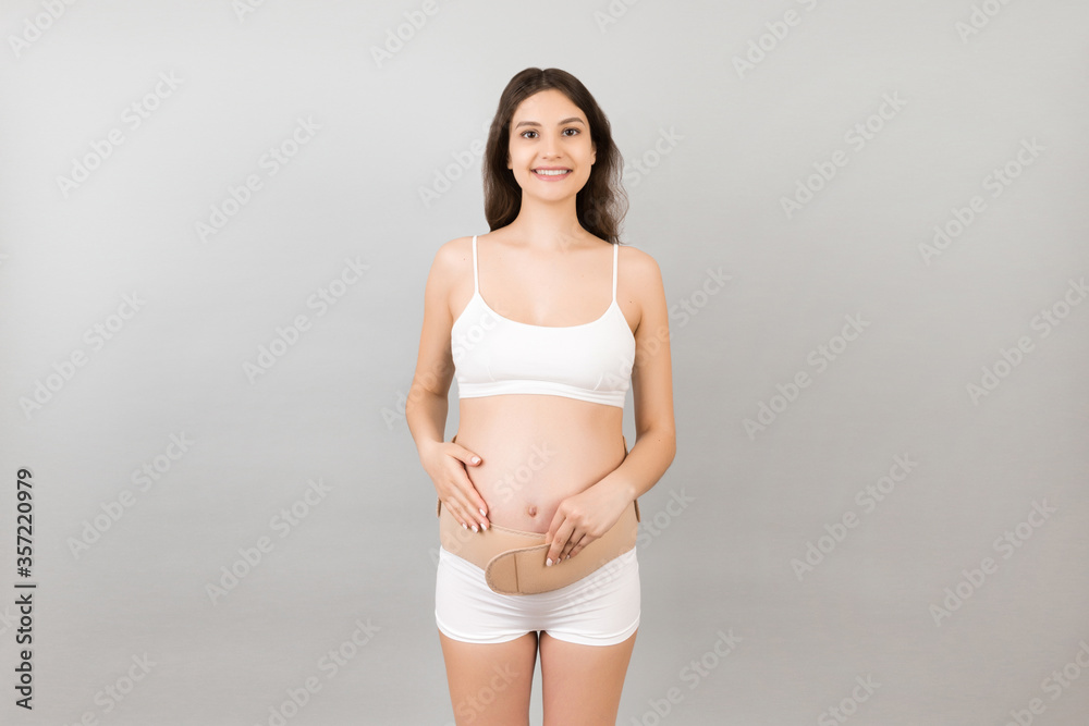 Close up of pregnant woman in underwear wearing elastic pregnancy bandage to make the pain go away at yellow background with copy space. Orthopedic abdominal support belt concept Close up of pregnant woman in underwear wearing elastic pregnancy bandage to make the pain go away at yellow background with copy space. Orthopedic abdominal support belt concept