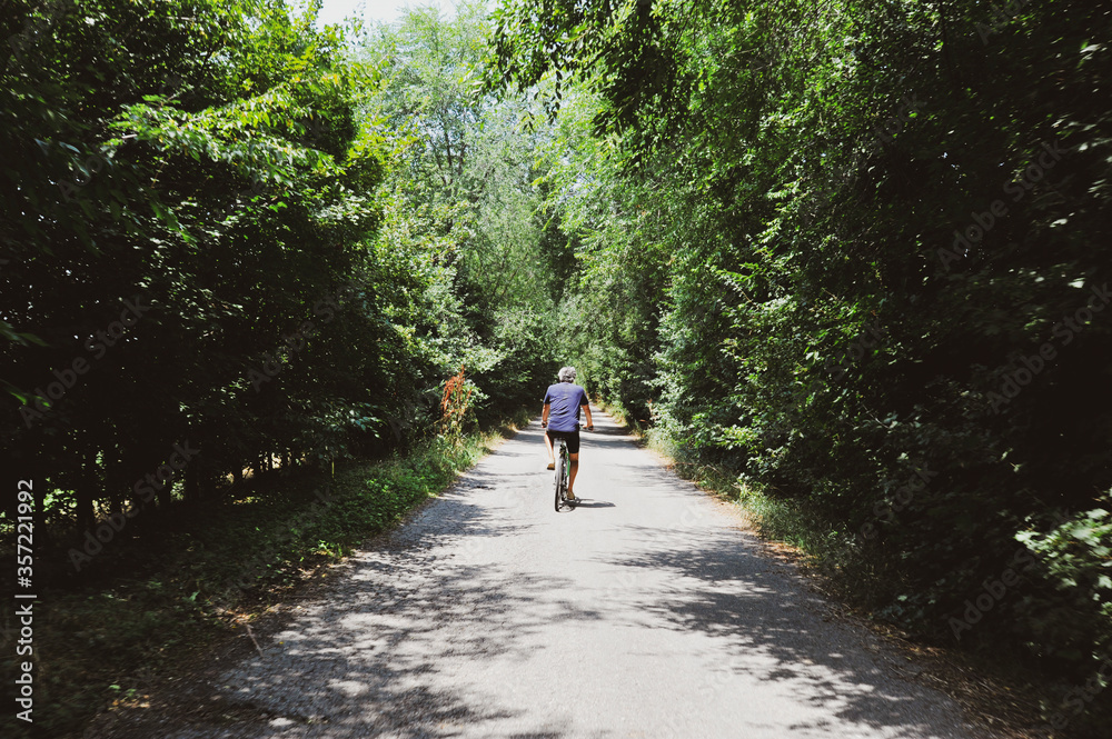 Rear view of active senior man riding bicycle along forest road on summer day - Senior athletic man cyclist riding bike on the trail in sunny forest - Healthy lifestyle and travel concept - Copy space