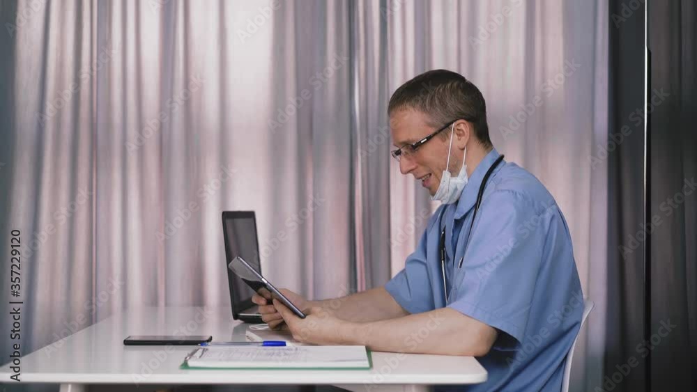 upset doctor wearing blue uniform and glasses with protective mask removed on neck does video call with tablet sitting at table in hospital office