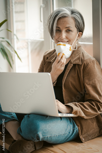 Grey haired woman removes mask affably looking at camera. Caucasian lady uses computer sitting tailor-fashion with crossed legs leaning back against window. Quarantine concept. Tinted image. photo