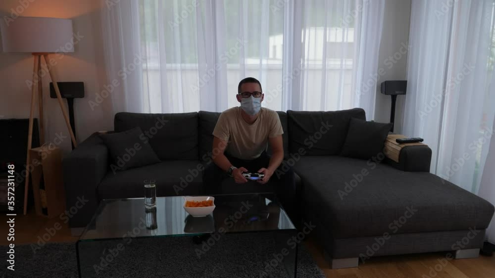 Young man with face mask sitting on sofa playing video games. Light from the television flickers, in the end happy about victory. Young man with face mask sitting on sofa playing video games. Light from the television flickers, in the end happy about victory.