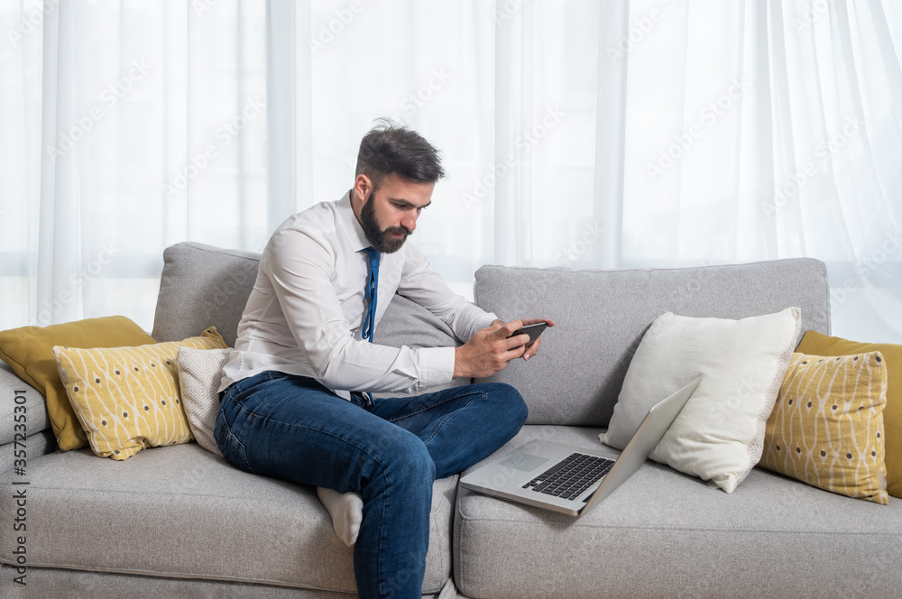 Young freelancer businessman sitting on sofa at his home and taking a photo with phone of laptop to show his boss charts he made for new business project as freelance worker 