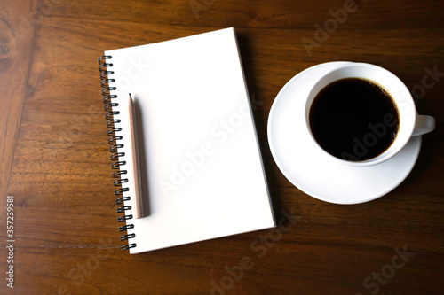 A white cup of hot Americano coffee with saucer and notebook on wooden table.Top view of open spiral blank notebook with wood pencil.