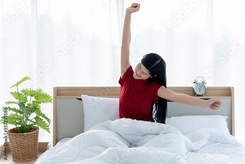 Pretty Asian woman is sitting on a clean white bed, stretching her arms to muscle relaxation After she woke up