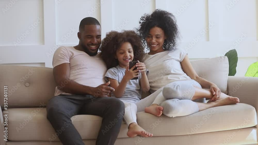 Happy african family with small cute kid daughter sit on sofa having fun with smart phone, little child girl holding cellphone using funny mobile app laughing with black parents take selfie at home Happy african family with small cute kid daughter sit on sofa having fun with smart phone, little child girl holding cellphone using funny mobile app laughing with black parents take selfie at home