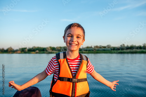 Happy smiling boy in orange life jacket by the lake
