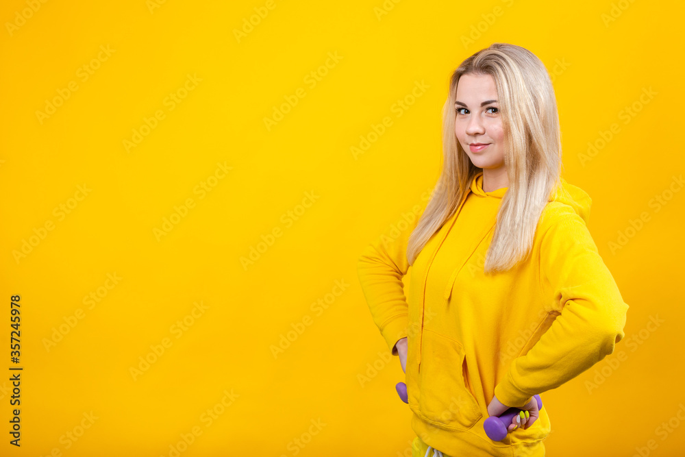 Happy beautiful young blonde woman in casual yellow sporty clothes doing exercises with small 0.5 kg violet dumbbells.