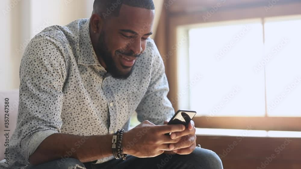 Young african american man holding smartphone device texting sms message sitting at home office, smiling black guy using apps playing mobile game chatting in social media surfing web on phone indoors