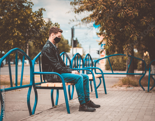 Caucasian  men sits on the bench and looks straight away. He is depressed. Dressed into jeans, T-shirt, leather jacket and black protective mask.  (Picture has a preset dark colors.) © Евгения Жигалкина