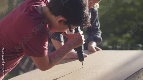 A father watches his teenage son driving a screw into a piece of wood with a power drill