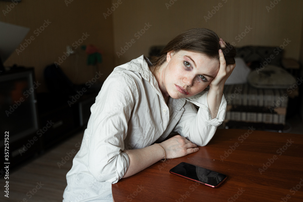 the victim of domestic violence and abuse, a girl sitting at a table, looking at the window holding her head in her hand, a man sleeping in the background