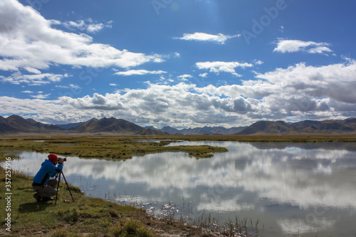 A tourist photographer setting up camera on tripod and taking a picture of beautiful reflection of hills and clouds in glass like waters of lake in Tibet, Asia