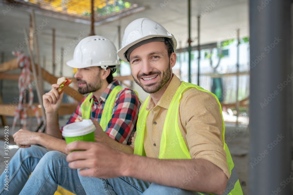 Building workers in yellow vests and helmets sitting on boards, having coffee with sandwiches, smiling Building workers in yellow vests and helmets sitting on boards, having coffee with sandwiches, smiling