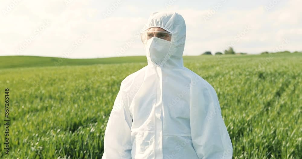 Portrait of Caucasian male farmer ecologist in white protective costume, mask and goggles standing in green field and turning face to camera. Man scientist and biologist posing in margin with harvest.