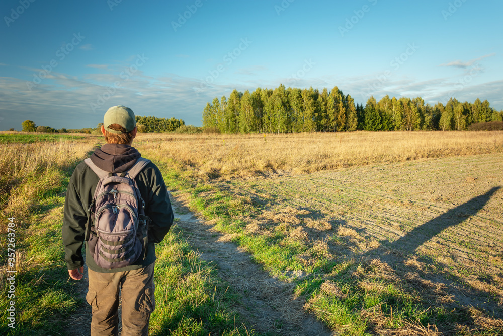 A man with a backpack walking dirt road through fields, forest and blue sky