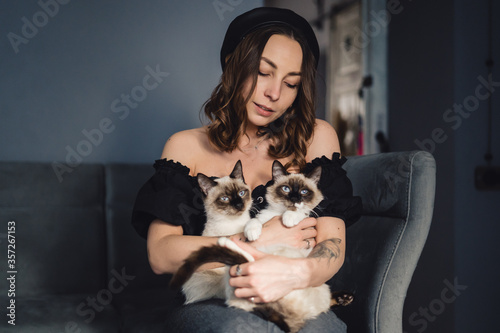 Attractive girl embracing her Siamese cats. Indoor portrait of cute woman playing with her two pets.