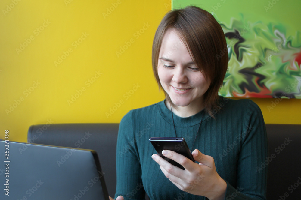 A laughing girl with a cheerful expression on her face and a smile sits at a laptop at a table in a cafe. She holds in her hand a smartphone, a mobile phone and looks at the screen of the gadget A laughing girl with a cheerful expression on her face and a smile sits at a laptop at a table in a cafe. She holds in her hand a smartphone, a mobile phone and looks at the screen of the gadget