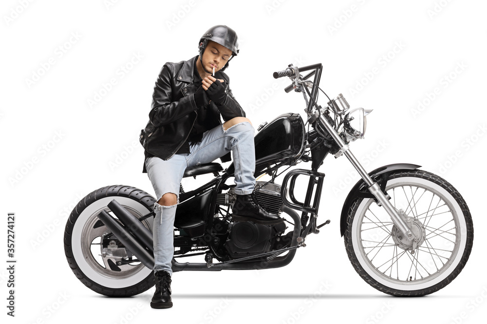Young biker with a helmet and a leather jacket sitting on a chopper motorbike and lighting a cigarette