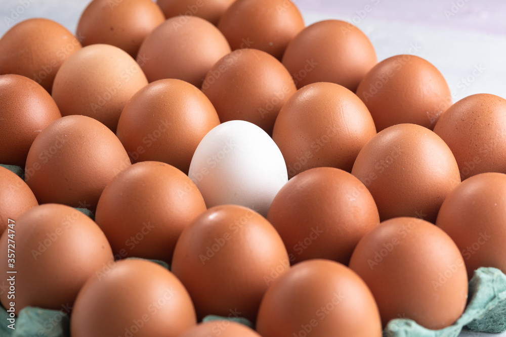 White egg stands out in a box of red eggs on a rustic white table. Top view.