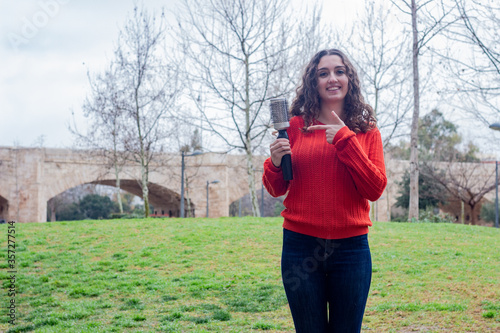 Portrait of pretty caucasian young woman showing rotating and styling brush  pointing the piece with index finger, in the park, orange sweater and jeans, long curly hair. Place for your text in copy s