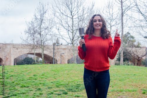 Portrait of attractive caucasian young woman model holding rotating and styling brush, keeping finger crossed, in the park, orange sweater and jeans, long curly hair. Place for your text in copy space