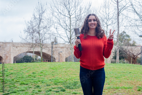 Portrait of young woman with rotating and styling brush  making victory or peace sign, in the park, orange sweater and jeans. Place for your text in copy space.