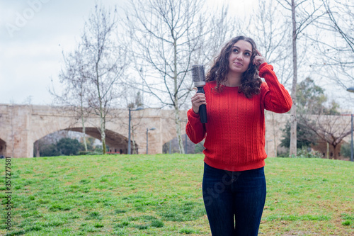 Portrait of attractive caucasian young woman model holding rotating and styling brush  thinks with finger on head, in the park, orange sweater and jeans, long curly hair. Place for your text in copy s