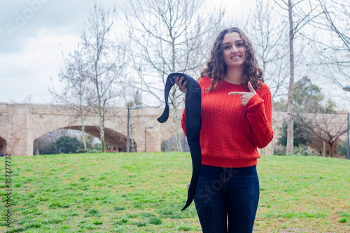 Portrait of pretty caucasian young woman model showing electronic abdominal appliance  pointing the piece with index finger, in the park, orange sweater and jeans, long curly hair. Place for your text