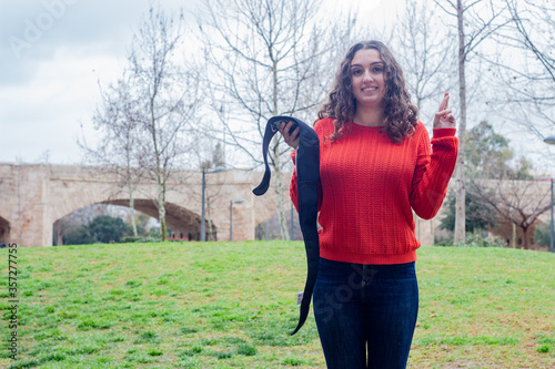 Portrait of attractive caucasian young woman holding electronic abdominal appliance, keeping finger crossed, in the park, orange sweater and jeans, long curly hair. Place for your text in copy space.
