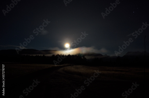 dreamy full moon night sky over field, forest road and mountain