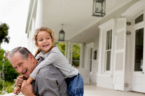 Slika na platnu Older man carrying granddaughter piggy back on porch