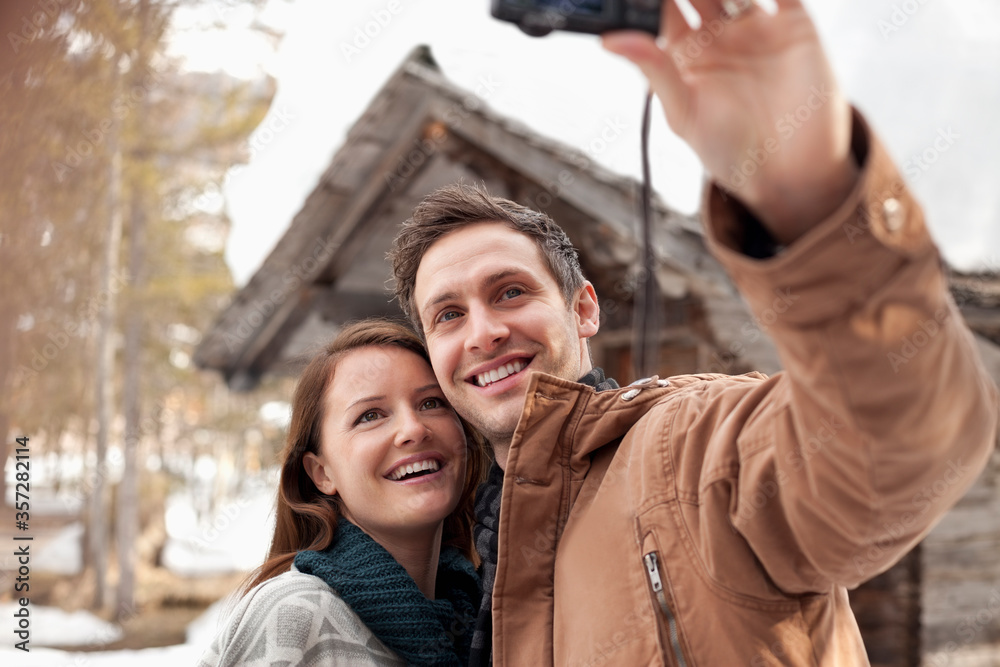 Smiling couple taking self-portrait digital camera outside snowy cabin